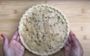 Hands holding unbaked chocolate chip cookie pie on wooden surface. Ready for oven baking.